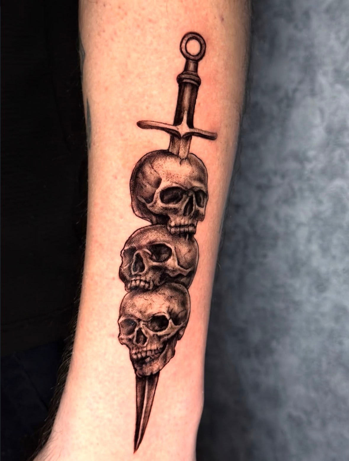 Black and grey realism tattoo of three stacked skulls pierced by a dagger on the forearm, El Artista Beirut